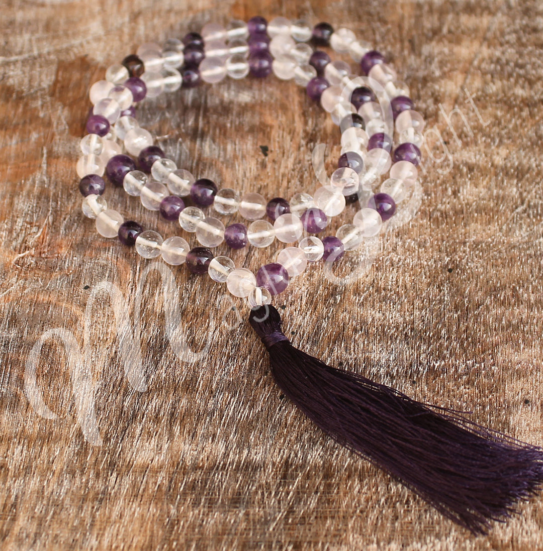 An amethyst, crystal quartz and rose quartz mala necklace with polished round beads and a long black tassel, displayed coiled on a wooden surface.