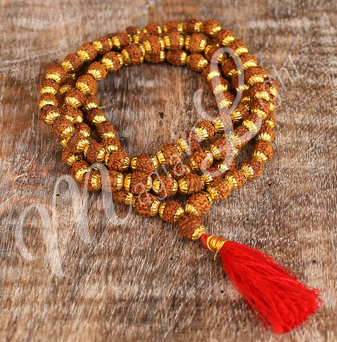 A rudraksha mala necklace with polished round beads with golden cap and a long red tassel, displayed coiled on a wooden surface.