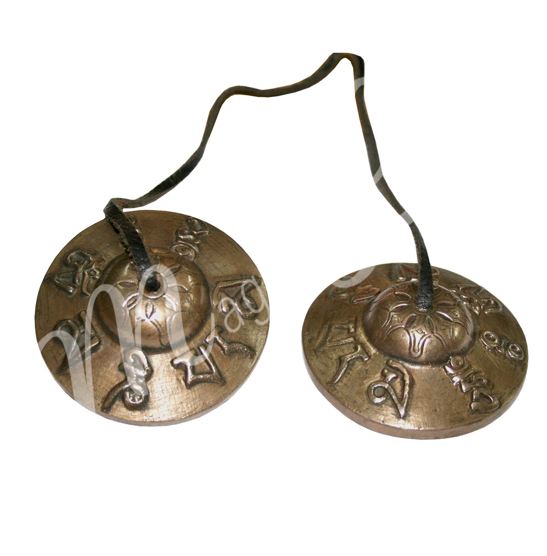 A pair of Tibetan tingsha cymbals connected by a leather cord, featuring engraved mantra symbols around the edges.
