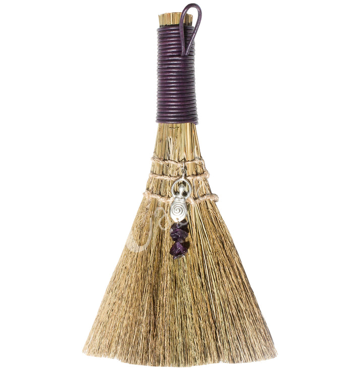A witch's broom with a purple leather cord on top, featuring a straw base, an Goddess and Amethyst beads.