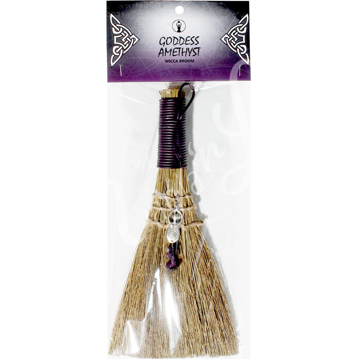 A witch's broom with a purple leather cord on top, featuring a straw base, an Goddess and Amethyst beads. In a retail bag with the words: Goddess Amethyst, wicca broom; on the card.