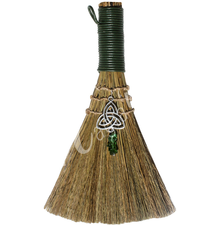 A witch's broom with a green leather cord on top, featuring a straw base decorated with a triquetra symbol and green Aventurine chips.