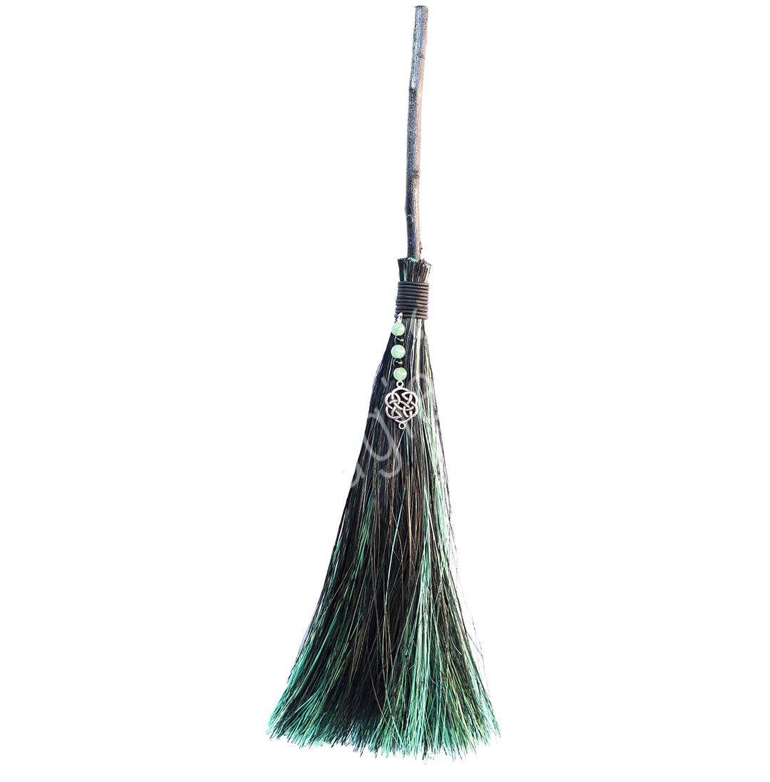 A witch's broom with a black and green head, green aventurine beads and a celtic knots charm. The broom has a metal handle.