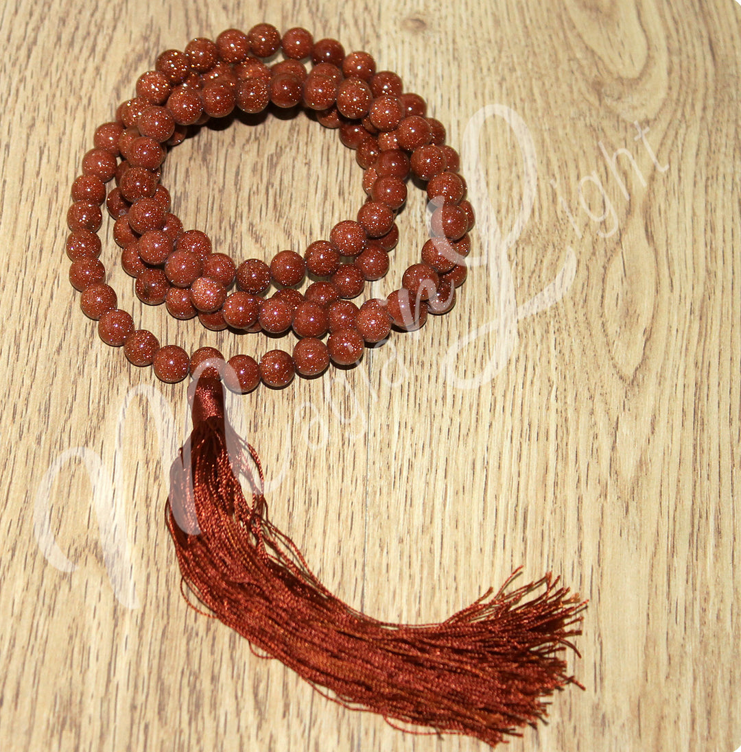 A goldstone mala necklace with polished round beads and a long red tassel, displayed coiled on a wooden surface.