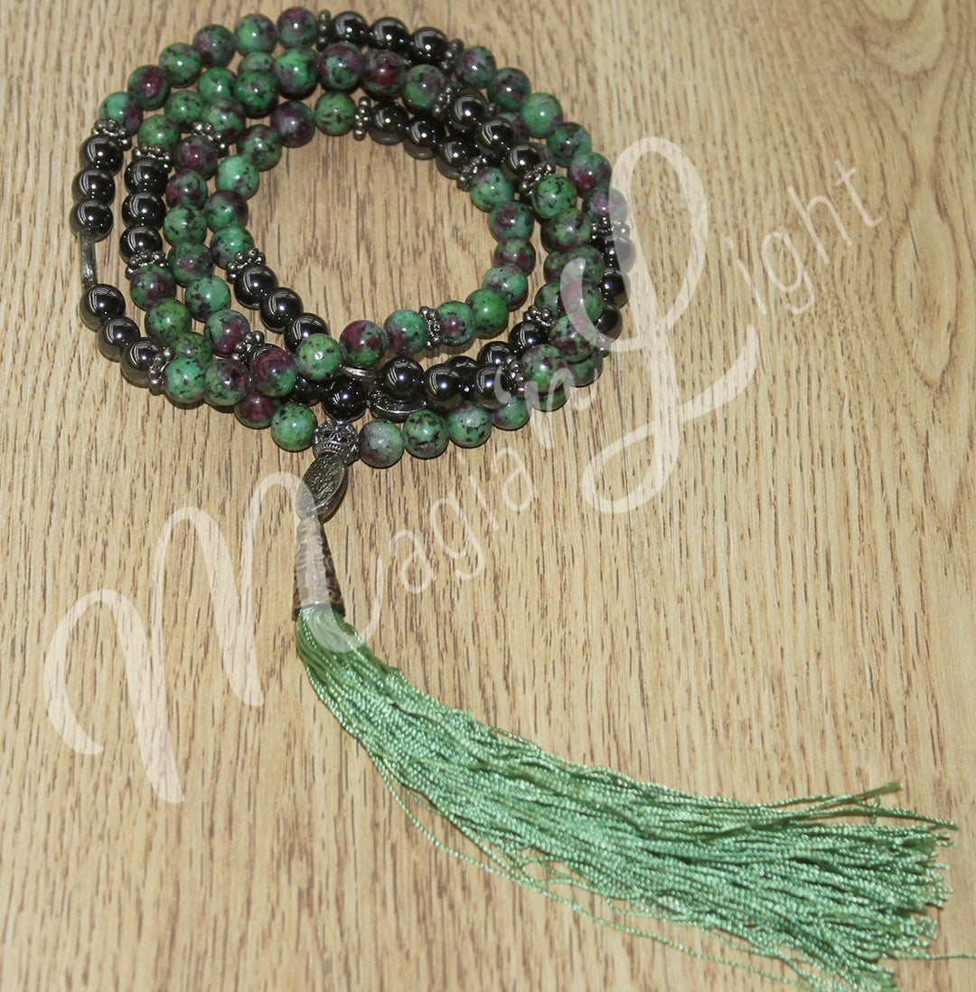 A ruby zoisite mala necklace with polished round beads with spacer beads and a long green tassel, displayed coiled on a wooden surface.