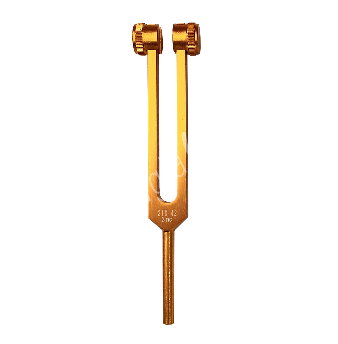 A orange tuning fork with the words "Sacral" engraved on it. It has two prongs with adjustable weights at the top, which are used to modify the vibration frequency.