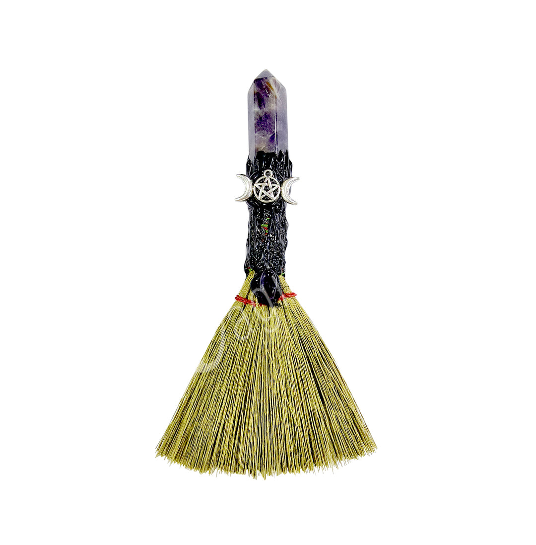 A decorative witch's broom with a pentacle in a triple moon charm attached to the handle, made from a amethyst point and a bundle of straws for the broom.