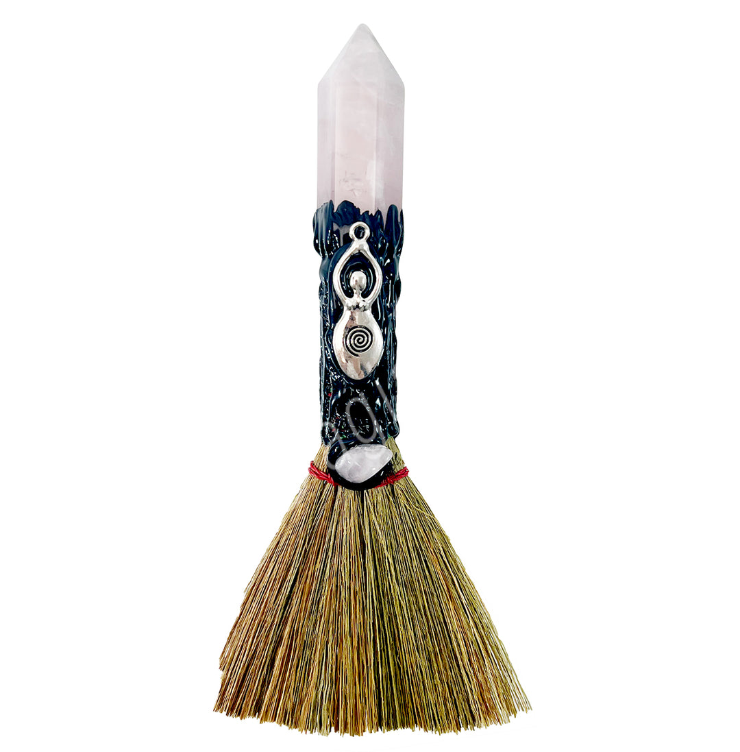 A decorative witch's broom with a goddess charm attached to the handle, made from a rose quartz point and a bundle of straws for the broom.