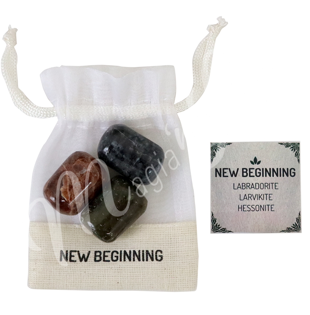 A white drawstring pouch labeled “NEW BEGINNING” displayed with three polished stones—Labradorite, Larvikite and Hessonite—alongside a tag that reads “NEW BEGINNING: Labradorite, Larvikite, Hessonite.”